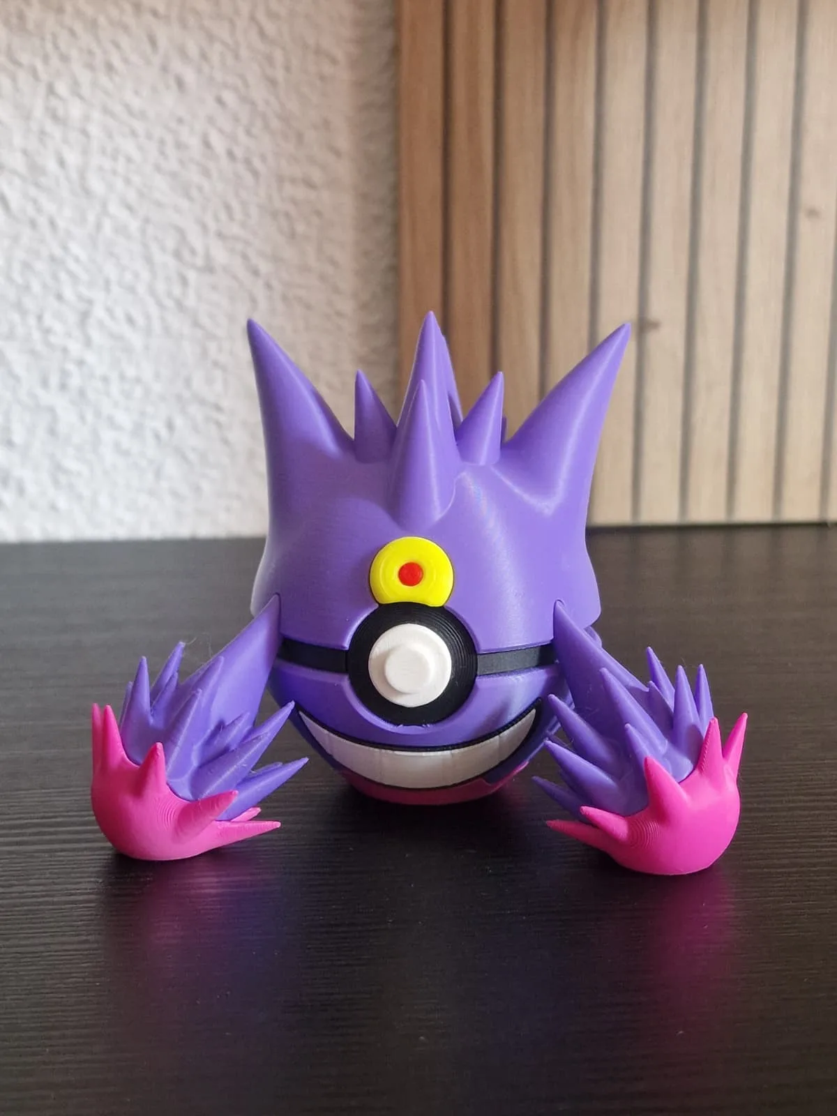 Mega Gengar saliendo de una pokeball: cuerpo morado con púas, garras rosa flúor apoyadas en la mesa y la bola blanca y negra en el centro