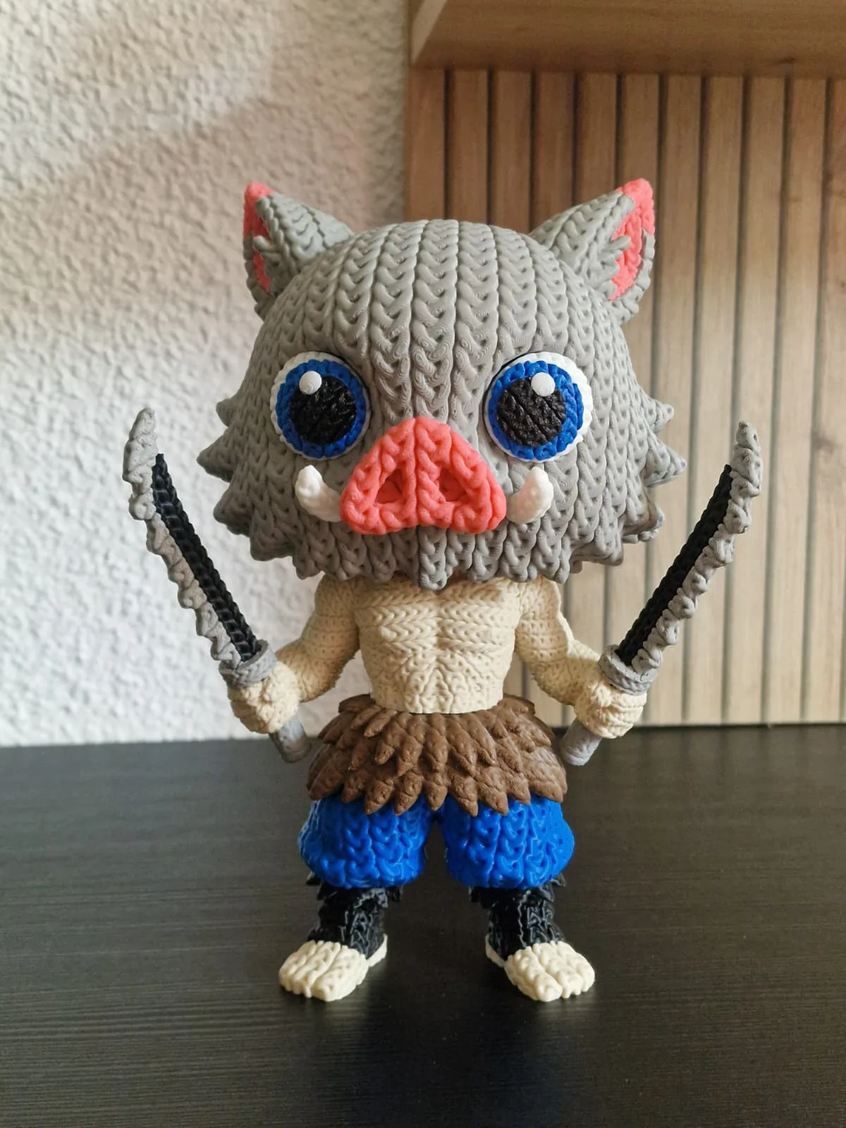 Figura de Inosuke con acabado de punto de ganchillo: máscara de jabalí gris con ojos azules y hocico rosa, dos katanas dentadas y pantalón azul