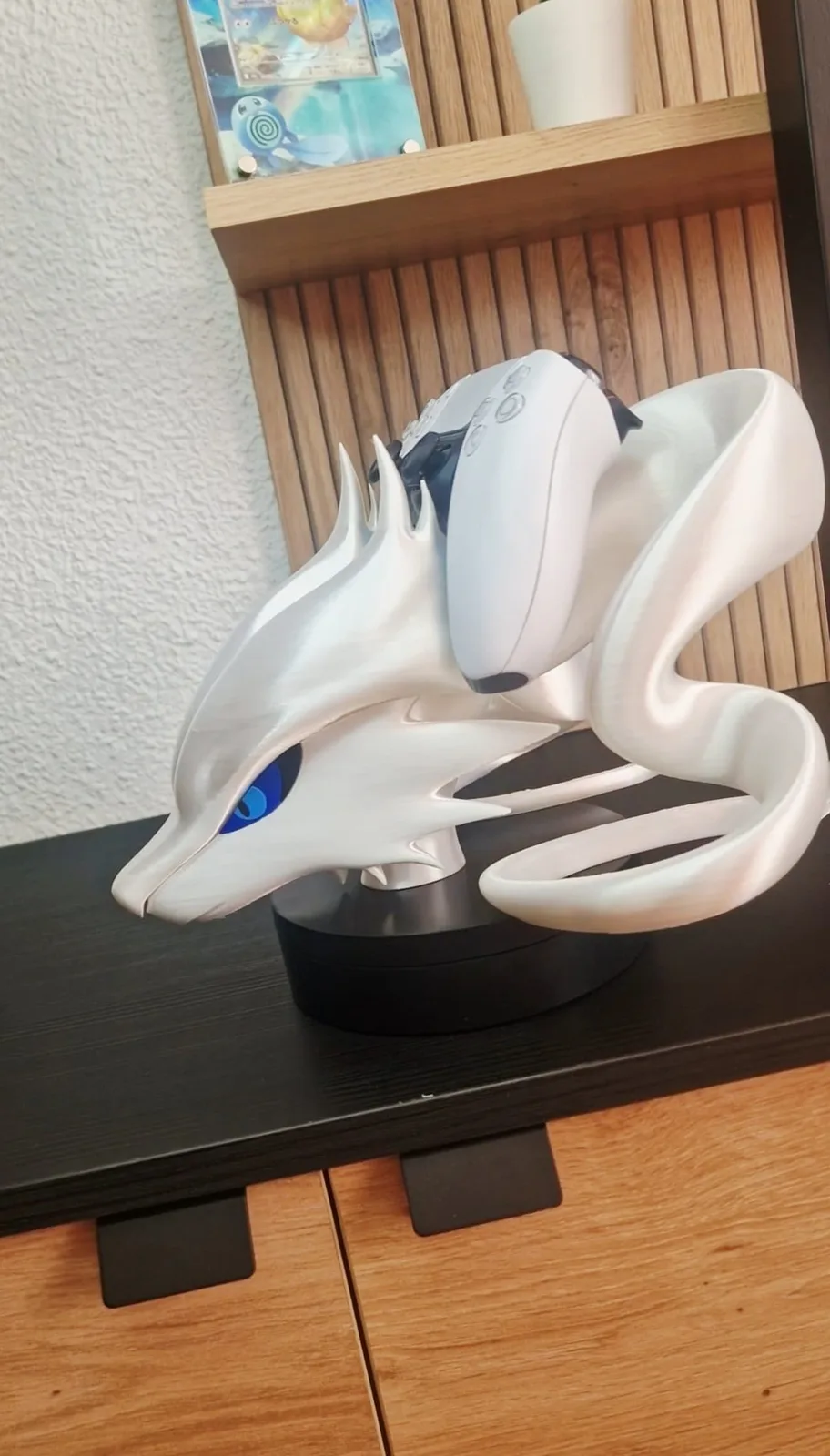 Soporte de mando Reshiram en blanco con los ojos azules, sujetando un mando DualSense blanco sobre una peana redonda negra